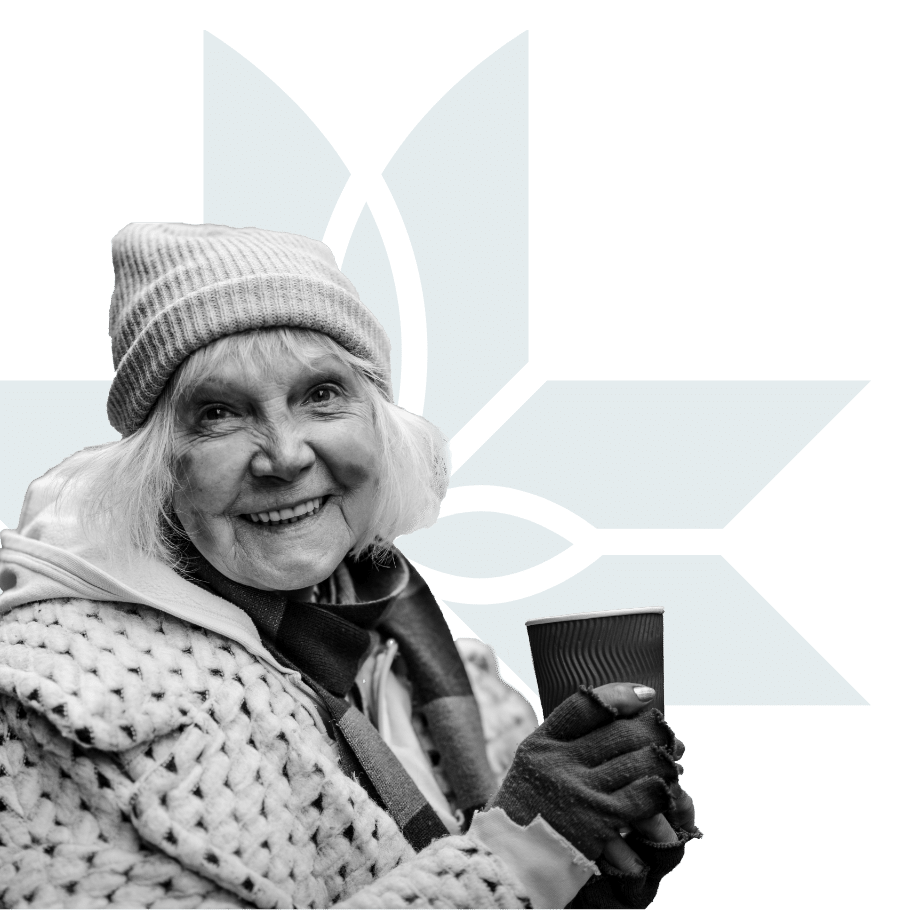 An elderly woman in a knit hat and coat smiles while holding a paper cup, standing in front of a blue geometric floral design. The image is black and white except for the blue background pattern.