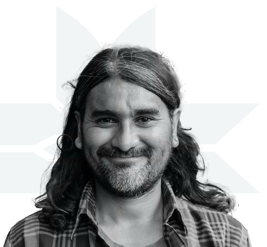A man with long hair and a beard smiles at the camera. He is wearing a plaid shirt. The background is white with a faint geometric pattern.