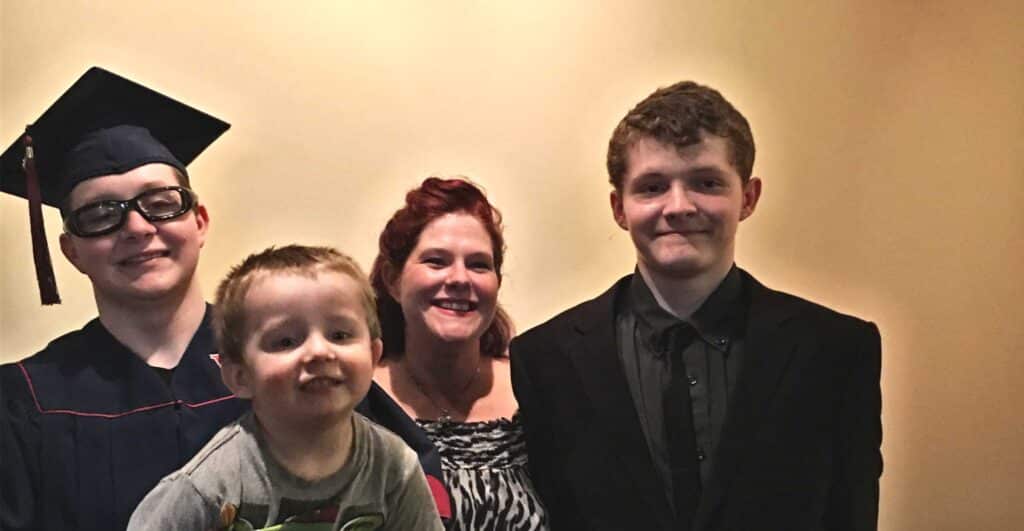 A woman smiles with three boys; one in a graduation cap and gown, one in a suit, and a young child in front. They stand against a plain beige background.